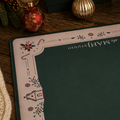 A square desk mat with a festive Christmas border design and the text 'the MAH STUDIO' is displayed on a wooden desk surrounded by holiday decorations, including a matching green box, ornaments, candles, and wrapped gifts.