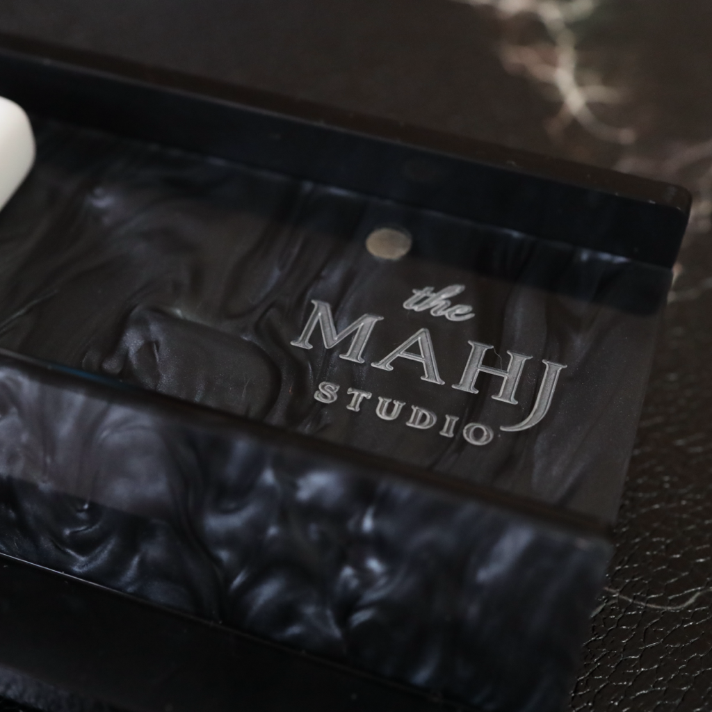 A close-up of a black textured acrylic or glass tray engraved with 'the MAHJ STUDIO' in silver lettering, resting on a dark marble surface next to a white object.