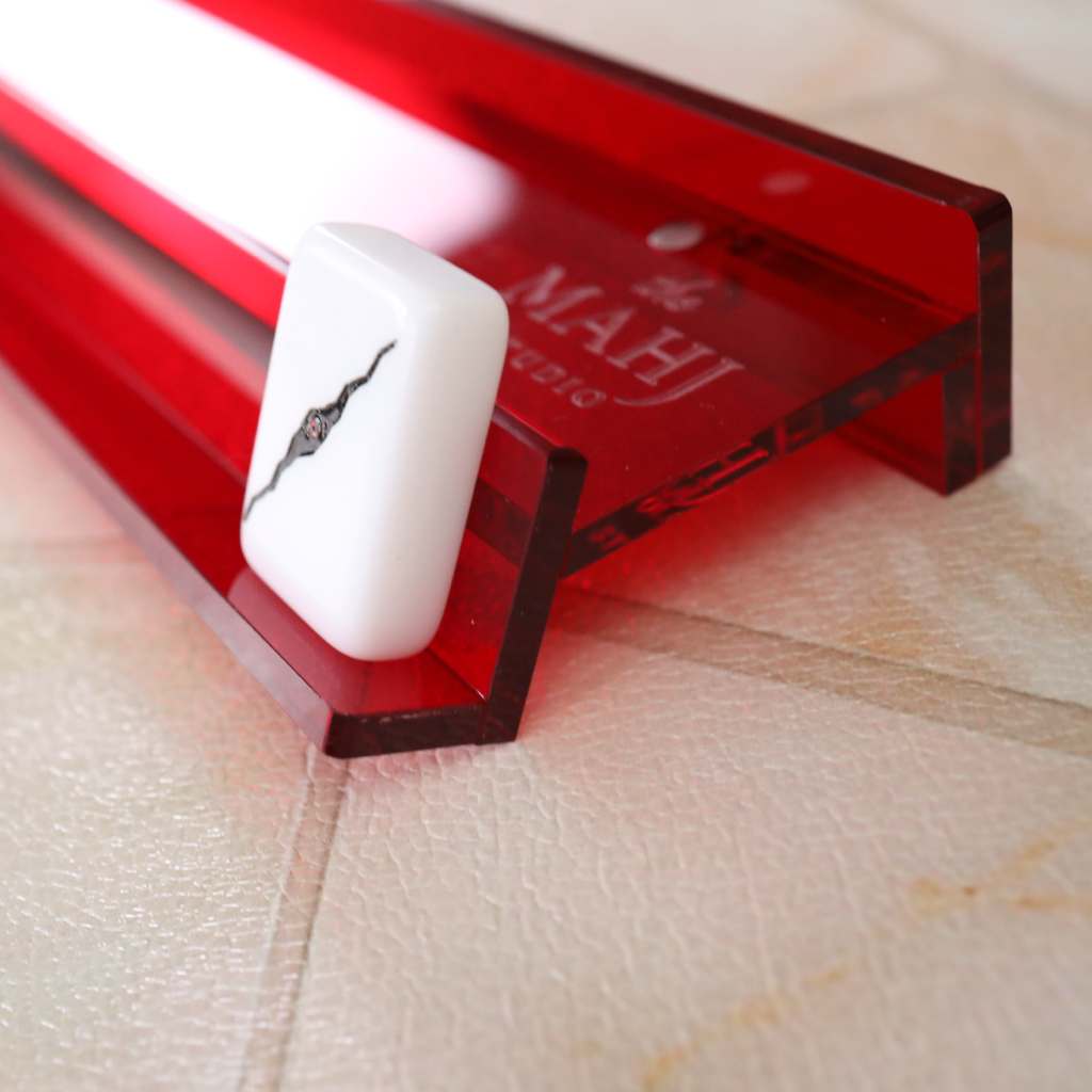 A close-up of a white rectangular product with a black crack design resting on a translucent red acrylic stand, set on a beige textured surface. The red stand has the brand name 'MAHI' printed on it.