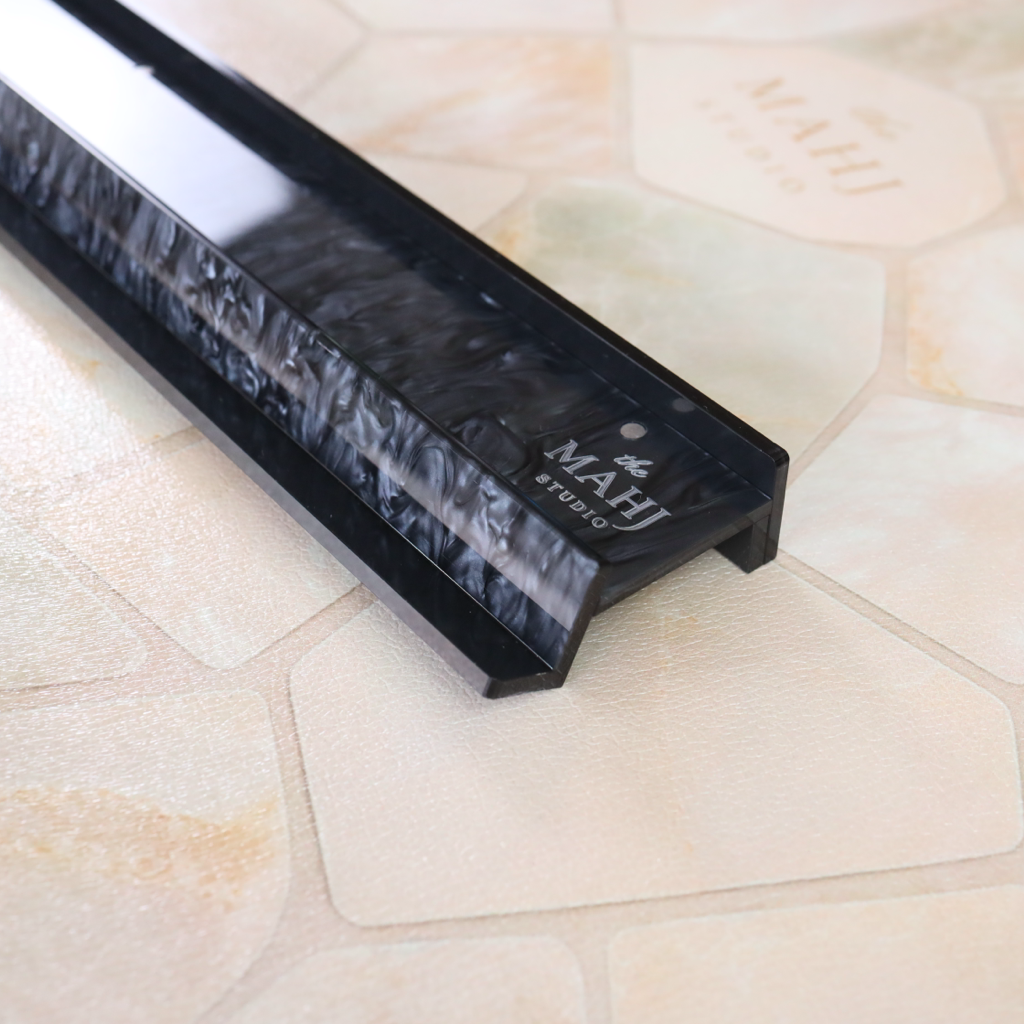 A close-up of a black marble-look linear channel or trim piece with the brand name 'MAHJ STUDIO' printed on it, resting on a beige and cream stone-look tile surface.