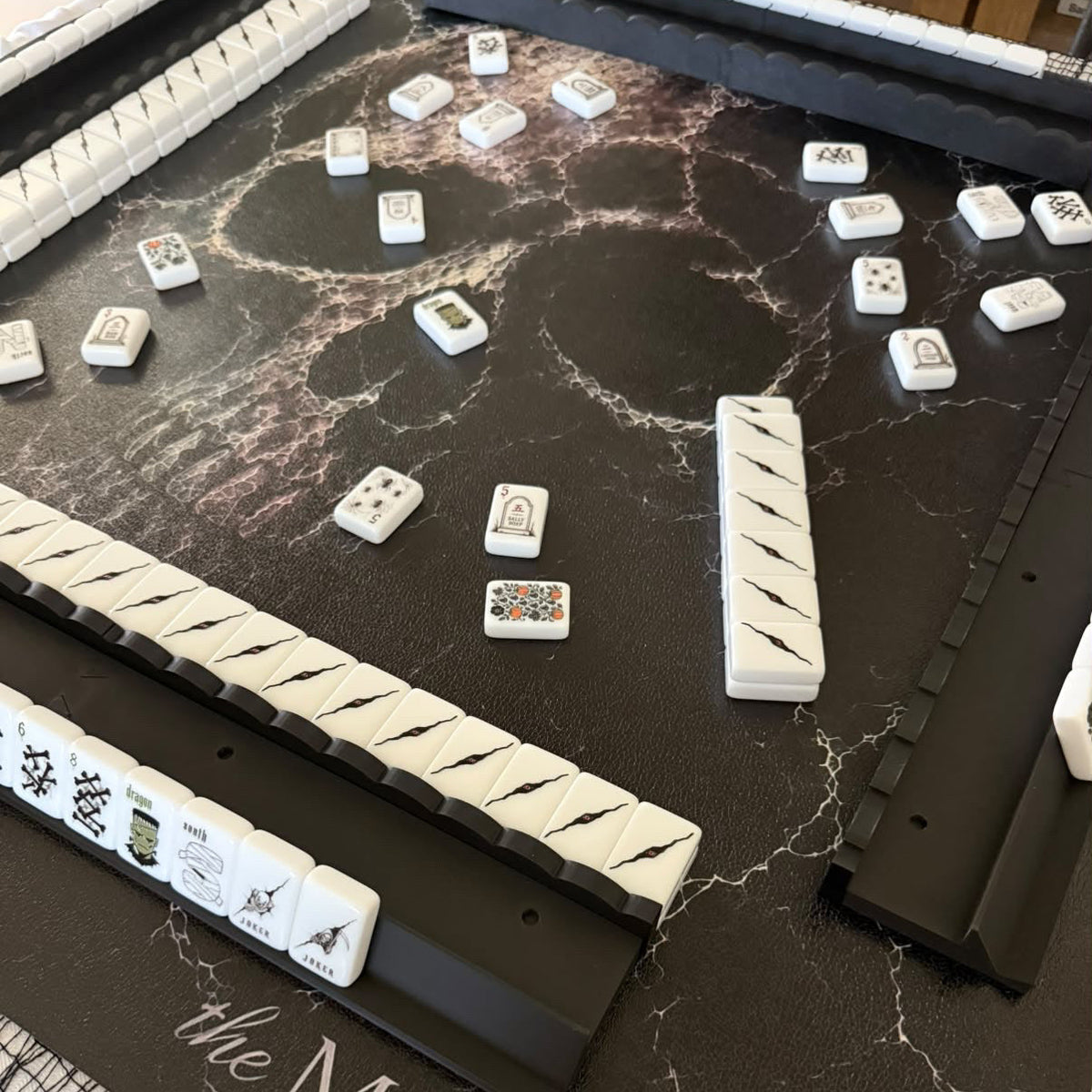 Mahjong game setup with multiple white and black tiles arranged on a dark marble-patterned mat. The mat has the text 'the MAHJ STUDIO' printed on it, and several racks of tiles are visible around the playing surface.
