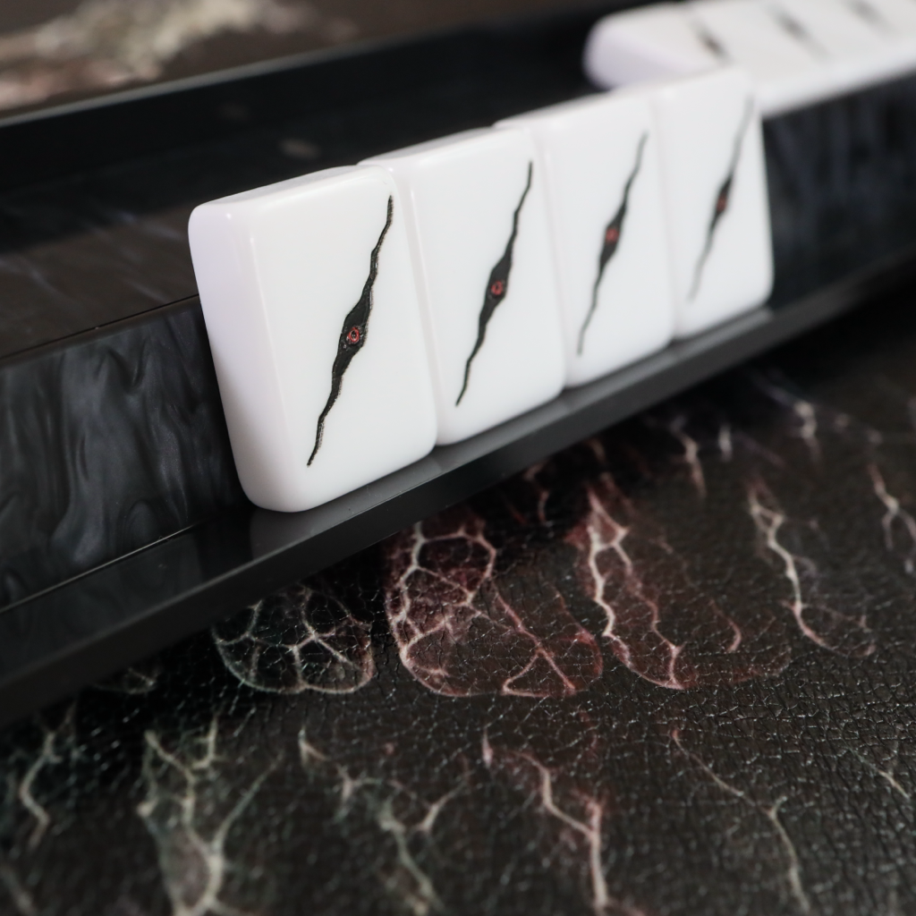 White mahjong tiles with black crack-like markings and a small red center detail, arranged on a glossy black rack over a dark marble surface.