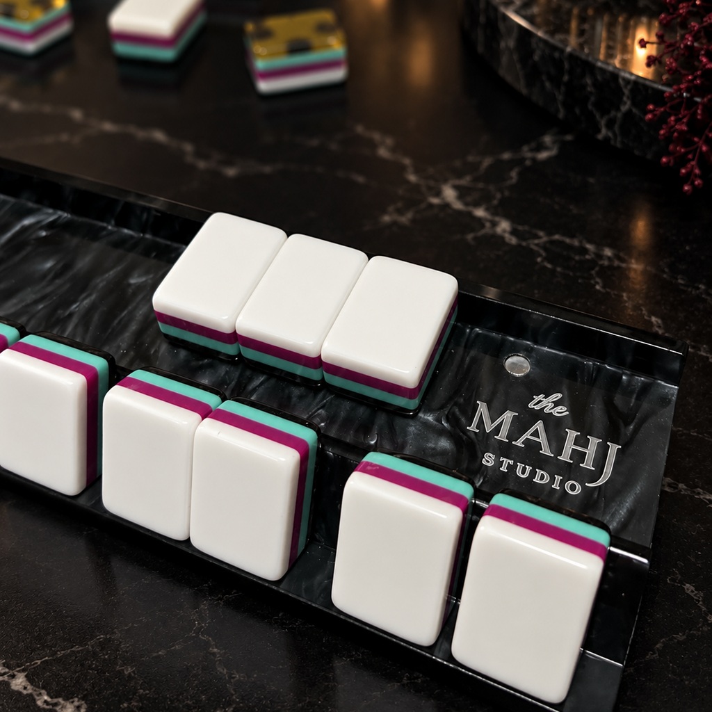 A close-up of a mahjong set on a black marble tray, with white tiles showing pink and teal edges. The tray is branded 'the MAHJ STUDIO'.