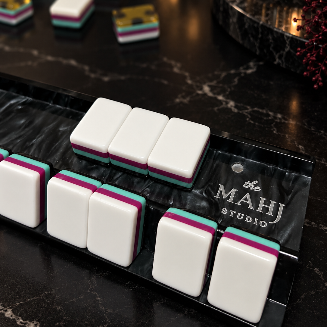 A close-up of a mahjong set on a black marble tray, with white tiles showing pink and teal edges. The tray is branded 'the MAHJ STUDIO'.