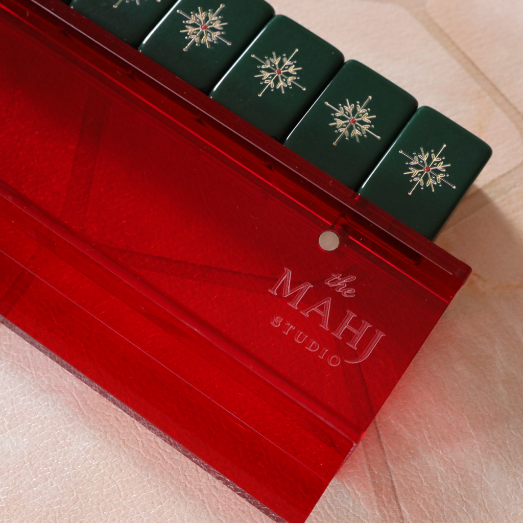 A close-up of a red acrylic mahjong rack holding dark green tiles with gold compass designs, placed on a light beige tiled surface. The rack is branded with 'the MAHJ STUDIO'.
