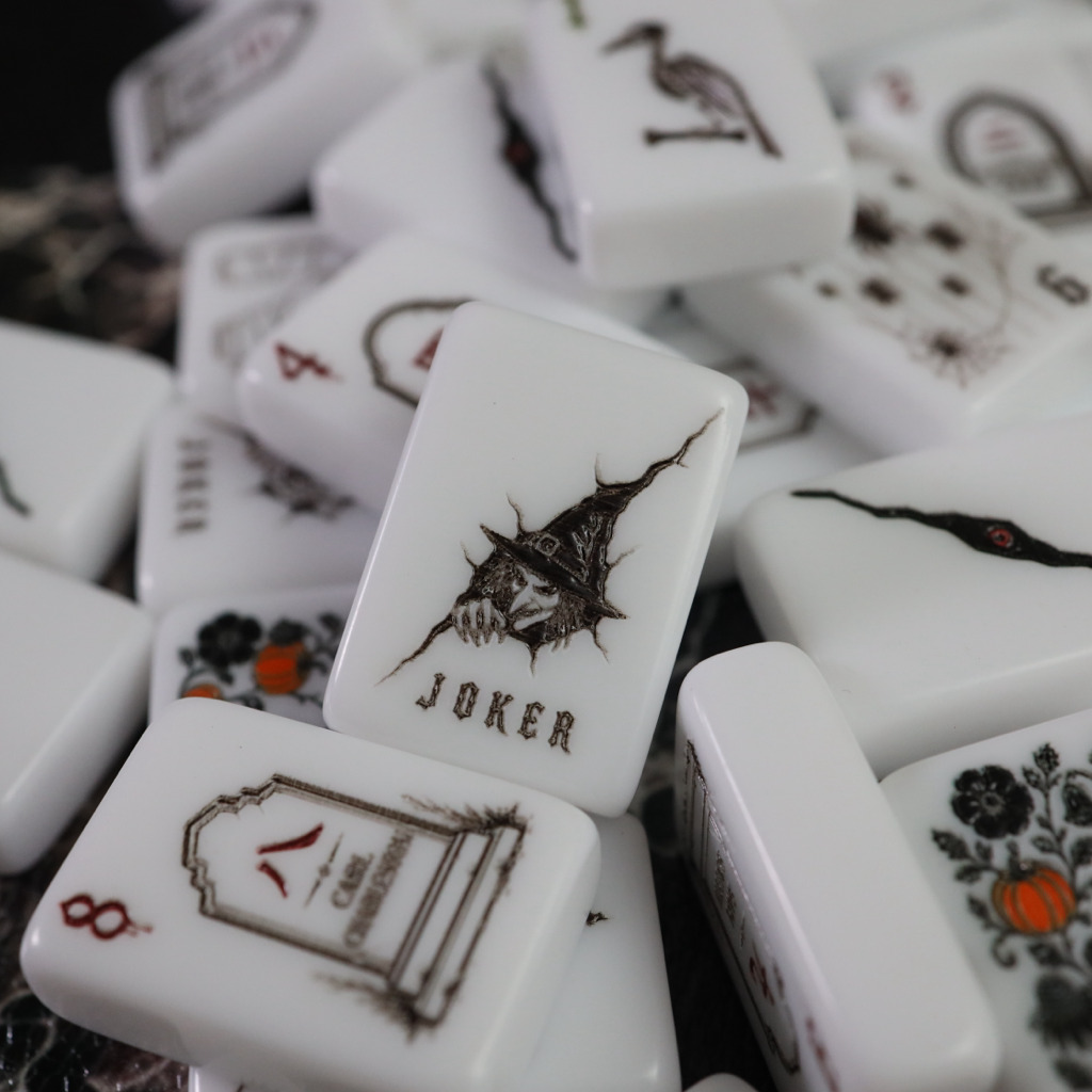 A pile of white ceramic mahjong tiles with black illustrations and symbols, including a tombstone marked 8 and a joker tile with a thorny design.