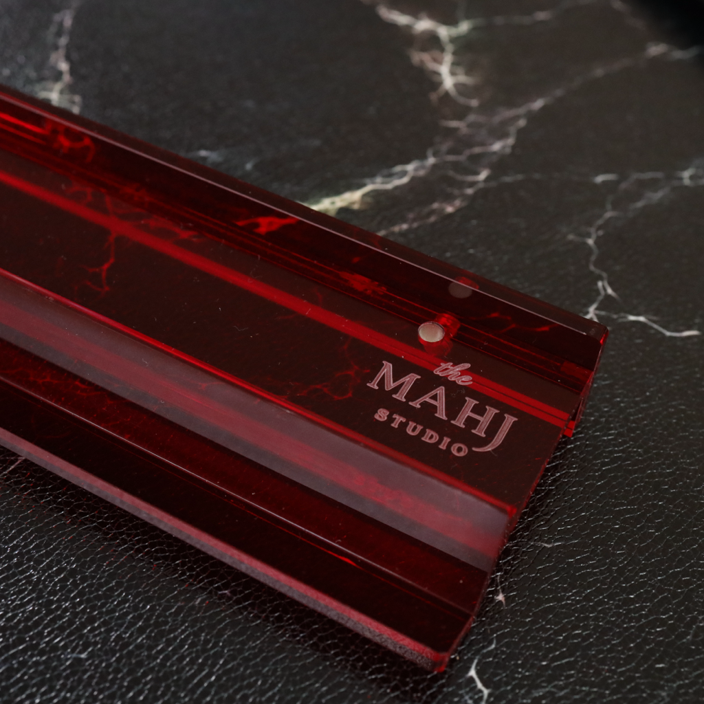 A close-up of a translucent deep red acrylic or glass ruler with the brand name 'MAHJ STUDIO' printed on it, resting on a black marble surface.