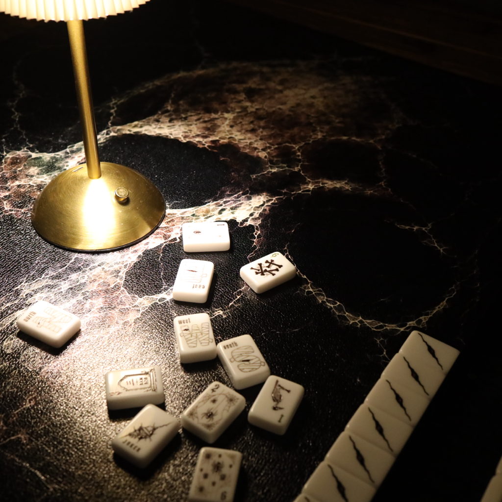 A set of white mahjong tiles with black markings arranged on a dark, glossy marble-patterned surface under the warm glow of a brass table lamp.