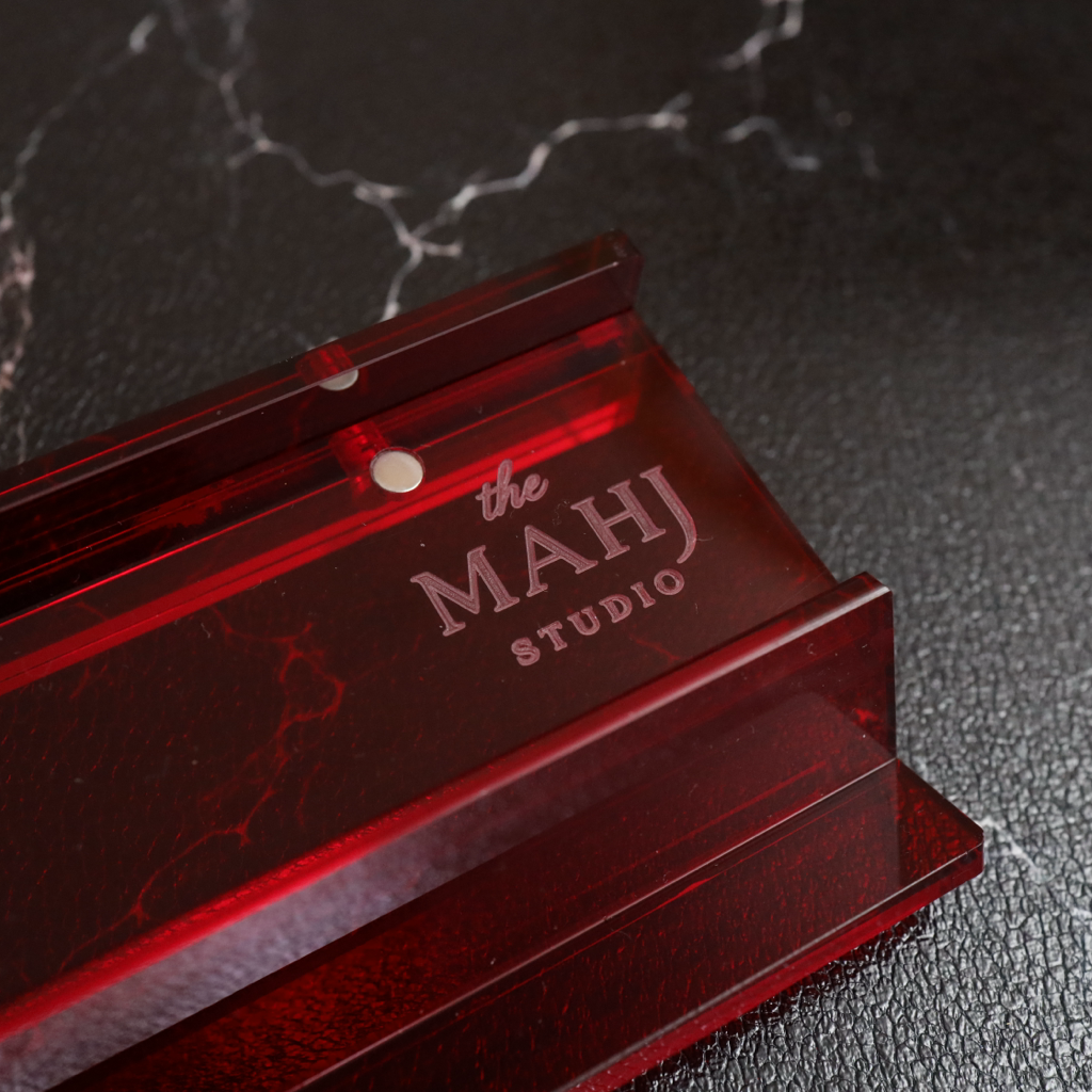 A close-up of a translucent dark red acrylic desk mat with 'the MAHJ STUDIO' logo printed on it, resting on a dark textured surface with a marble pattern.