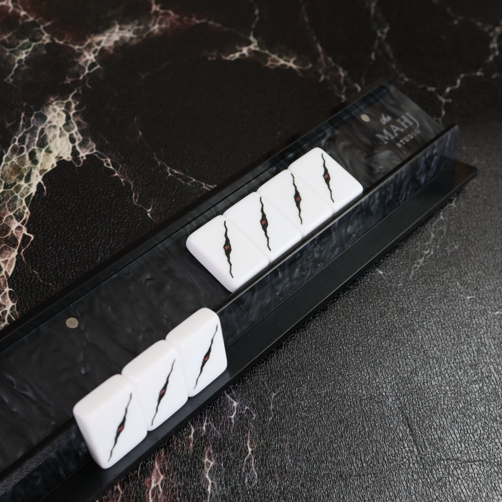 A close-up of a black mahjong rack holding several white tiles with black crack-like markings, placed on a dark marble-patterned surface.