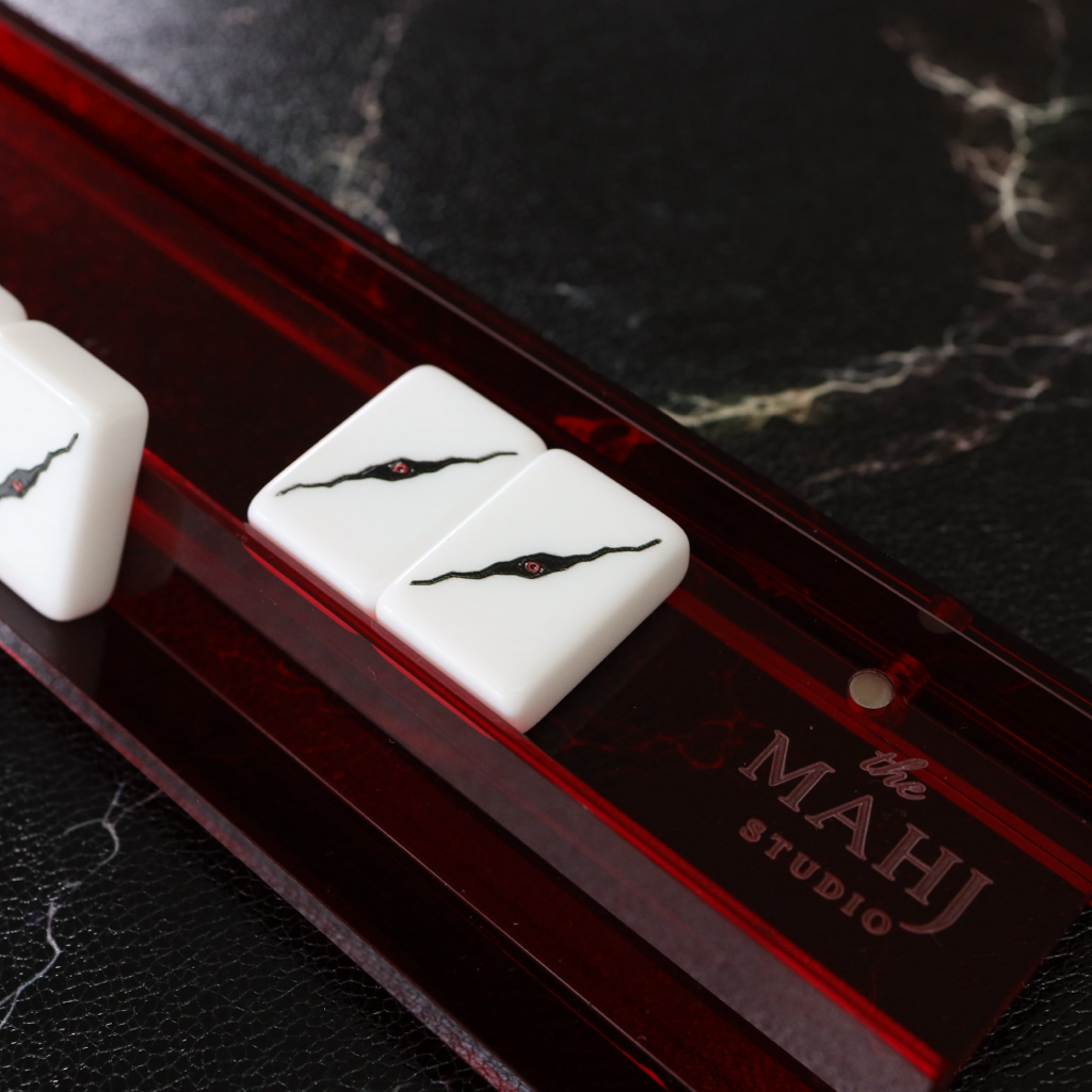 White mahjong tiles with black and red markings resting on a translucent dark red acrylic game board, photographed on a black marble surface. The board is branded with 'The MAHJ STUDIO'.