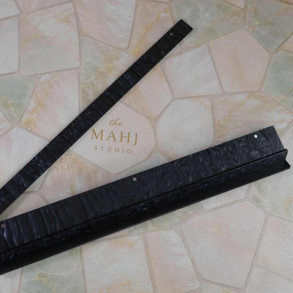 Two long black textured strips, likely hair extensions or trimmer guards, laid diagonally on a beige stone tile floor with 'the MAHJ STUDIO' logo visible in the background.
