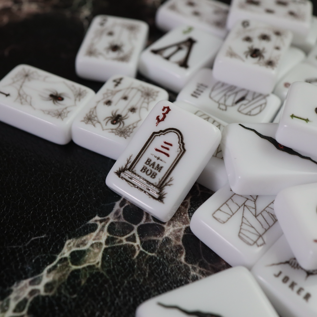 White mahjong tiles with black and red gothic-themed illustrations, including tombstones and spider motifs, scattered on a dark textured surface.