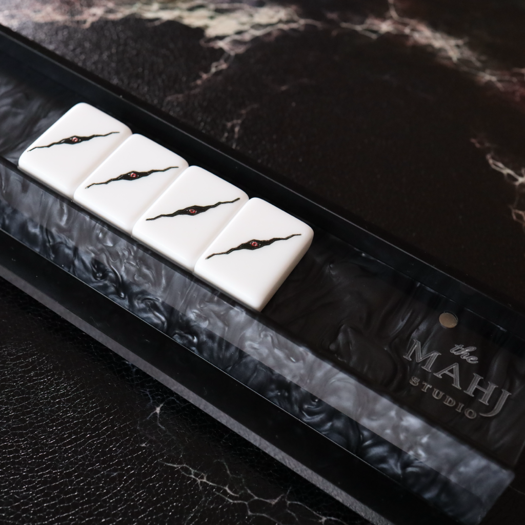 A close-up of a mahjong set showing four white tiles with black crackle patterns resting in a black and white marbled holder, placed on a dark textured surface. The brand name 'The MAHJ STUDIO' is visible on the holder.