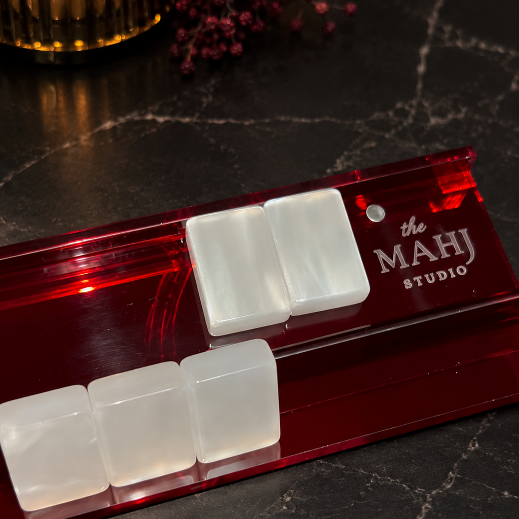 A red acrylic mahjong rack holding four white rectangular tiles, photographed on a dark marble surface with a candle and dried flowers in the background. The rack is branded 'the MAHJ STUDIO'.